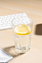 A clear glass filled with water and a lemon slice on a light wooden desk. Nearby, a white computer keyboard.Office environment, suggesting a refreshing break or an attempt to stay hydrated during work