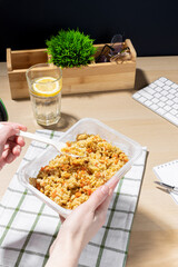 Office environment, break for nourishment. Snack at the workplace. Container of fried rice with carrots and chicken placed on a striped cloth. A hand holds a plastic fork. A glass of water with lemon.