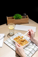 Office environment, break for nourishment. Snack at the workplace. Container of fried rice with carrots and chicken placed on a striped cloth. A hand holds a plastic fork. A glass of water with lemon.