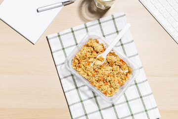 Office environment, break for nourishment. Snack at the workplace. Container of fried rice with carrots and chicken placed on a striped cloth. A glass of water with lemon. Nearby keyboard