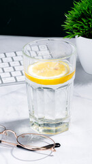 A clear glass filled with water and a lemon slice on a light desk. Nearby, a white computer keyboard.Office environment, suggesting a refreshing break or an attempt to stay hydrated during work