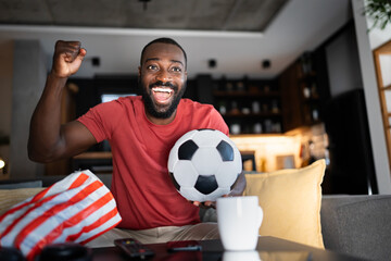 A cheerful man in a red shirt excitedly watches a soccer game at home, holding a soccer ball and...