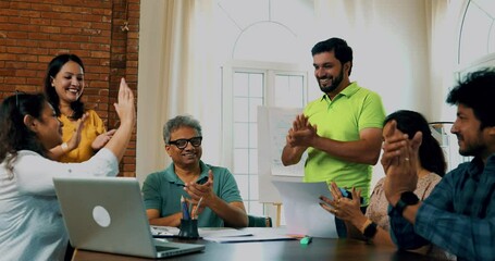 Indian Asian young business people or startup partners celebrating team success with high-fives and cheers while sitting in a meeting with a laptop and documents on the table in a modern cozy office