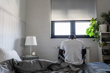 A man sits on the edge of his bed in a modern, minimalist bedroom, looking down in deep thought....