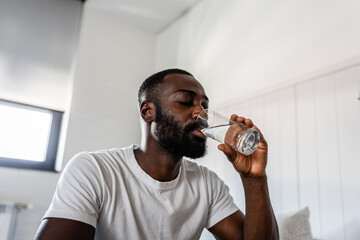A young man sits on his bed in the morning, drinking a glass of water. He looks refreshed and...