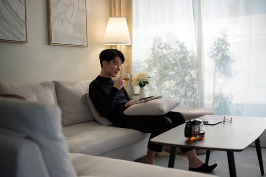 Calm young man in a black sweater sits comfortably on sofa, sipping a warm tea. - Powered by Adobe