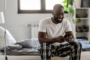A man in pajamas sits on his bed, smiling as he looks at his smartphone. The cozy bedroom setting, soft lighting, and relaxed posture convey a sense of comfort and enjoyment in a digital moment.