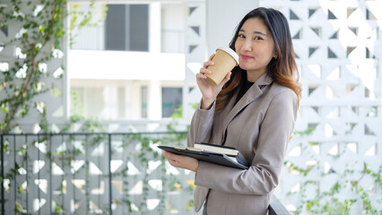 Fototapeta premium Smiling businesswoman in formal business attire sips coffee while standing against a contemporary backdrop of a stylish office building with greenery.