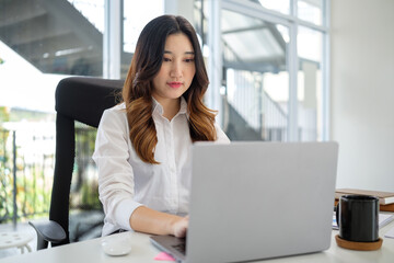Determined young woman in formal attire focuses on her laptop while seated at a sleek, modern desk.