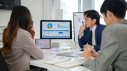 Obraz premium A group of professionals engaged in a strategic discussion while reviewing financial charts and graphs on computer screens in a bright, modern office setting.