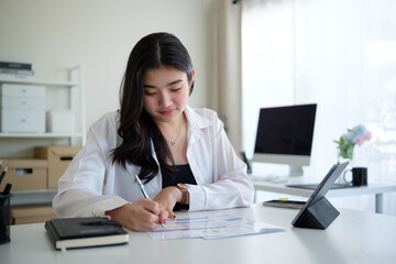 Focused businesswoman in a white shirt works on financial documents, taking notes in a modern and well-lit office.