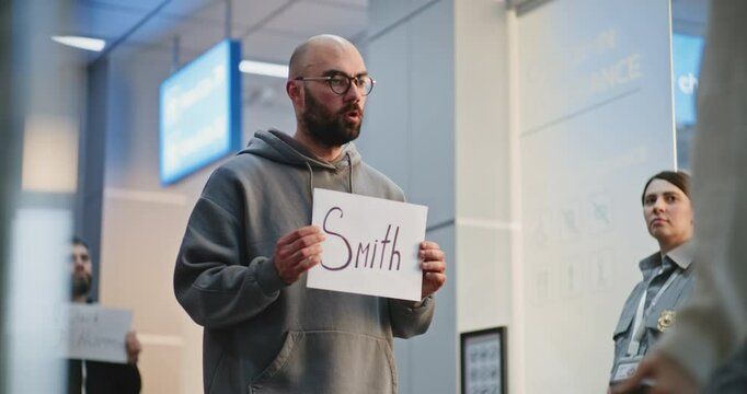 International Airport Terminal: Airport Greeter or Chauffeur Meeting Passengers From Flight with Name Sign. Diverse People Walking with Luggage. Business Transfer, Transportation Service. Medium Shot.
