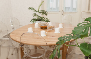 Cozy dining setting featuring a round wooden table surrounded by transparent chairs, complemented by lush green potted plants, creating a warm and inviting interior atmosphere.