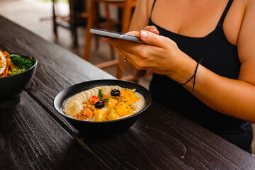 Woman has breakfast with delicious oatmeal with bananas, mangoes and berries, she takes pictures on mobile camera and posts it on social networks. Concept of counting calories using a mobile app