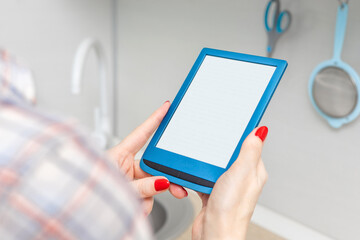 woman reading e-book in kitchen at home. housewife reading e-book. woman holding e-book in hand at home. ebook mockup. place for inscription. female hand with red manicure holding a book