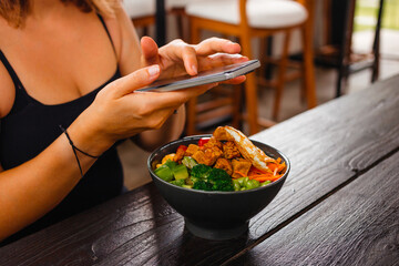 Woman takes a picture of a plate with salad mix, broccoli, edamame, tofu and a fried egg on her mobile phone. Concept of counting calories using a mobile application.