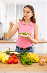 Vegeterian woman standing at table in kitchen and eating vegetable salad