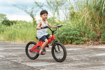 Young boy riding a red bicycle outdoors on a concrete path with tall grass and greenery in the background, showing an excited expression.