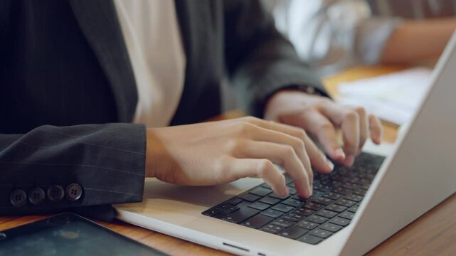 A woman is typing on a laptop computer. She is wearing a black jacket and white shirt. The laptop is open and the keyboard is visible. There is a tablet on the table next to the laptop