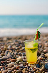 Fresh yellow cocktail with mint leaves resting on rocky shore near sparkling ocean waves symbolizes tropical refreshment and carefree vacation vibes