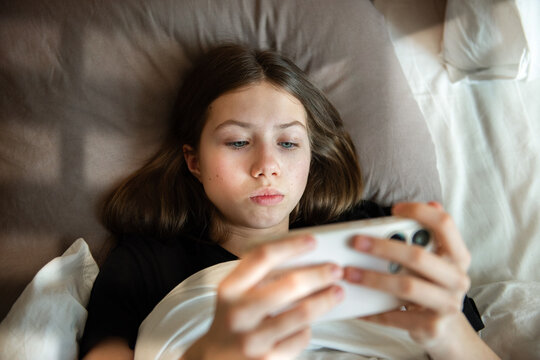 Teenage girl is lying in bed and holding a smartphone in her hands. Unaltered photo. Smartphone addiction and social media addiction.