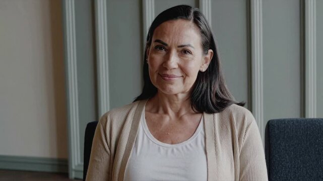 A brunette 50 year old woman in a simple cardigan sits and smiles in a waiting room chair, simple decor composition