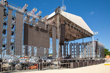Large outdoor concert stage under construction with LED screens, lighting rigs, and sound systems set up on a sandy beach under a clear blue sky...