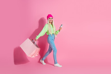 Stylish young blonde woman holding shopping bags and smartphone against pink background