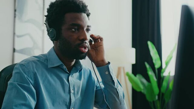 Focused African American man wearing headphones and blue shirt while sitting in modern room. Listening attentively. Adjusting headset while engaging in online meeting or attending virtual conference.