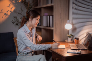 Young Asian businesswoman sitting at her desk working late at night in her home office taking notes on a notepad with laptop and calculator near, desk lamp illuminating workspace