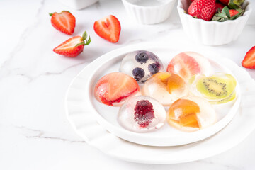 Japanese Raindrop Cake, or Mizu Shingen Mochi, clear jelly dessert, crystal-clear water-like delicacy agar pudding with fruit and berry slices, portioned on plates, white marble background copy space