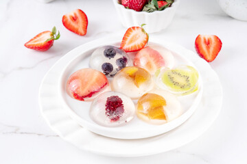 Japanese Raindrop Cake, or Mizu Shingen Mochi, clear jelly dessert, crystal-clear water-like delicacy agar pudding with fruit and berry slices, portioned on plates, white marble background copy space