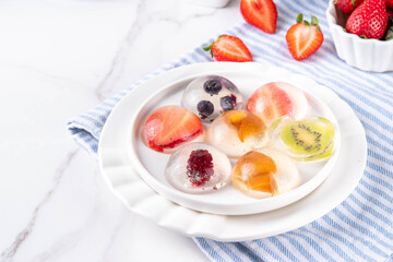 Japanese Raindrop Cake, or Mizu Shingen Mochi, clear jelly dessert, crystal-clear water-like delicacy agar pudding with fruit and berry slices, portioned on plates, white marble background copy space