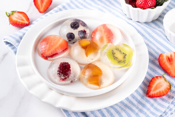 Japanese Raindrop Cake, or Mizu Shingen Mochi, clear jelly dessert, crystal-clear water-like delicacy agar pudding with fruit and berry slices, portioned on plates, white marble background copy space