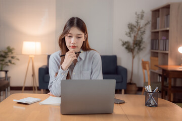Focused Asian businesswoman working remotely from home office using laptop computer and thinking with pen in hand, sitting at desk with calculator, notepad, smartphone and pen holder