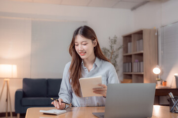 Young asian businesswoman calculating domestic expenses using calculator and taking notes in notebook while working with laptop computer at home office at night