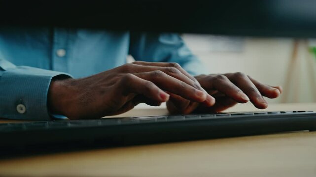Focused hands of African American man wearing blue shirt typing on keyboard while working at desk. Rapidly inputting data or composing message in professional setting with modern workspace elements.