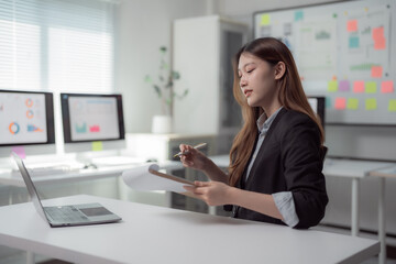 Young businesswoman reviewing documents and taking notes while working with financial data on laptop and desktop computers in bright modern office