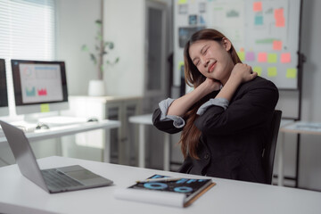 Obraz premium Asian businesswoman massaging her neck while seated at her desk, experiencing discomfort from neck pain after a long day of work in a busy office environment with a laptop