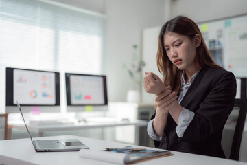 Asian female office worker is massaging her wrist, suffering from wrist pain after long time using computer, laptop, mouse while working in the office