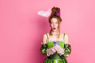 Fototapeta premium Young woman dressed in elegant green baroque gown holding phone against pink background, showing modern life in a historic comparison
