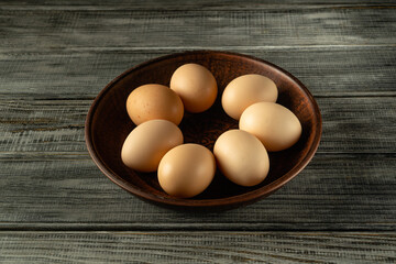 Fresh eggs of various shapes and shades are neatly placed in a dark wooden bowl resting on a textured wooden surface, showcasing a natural and organic feel