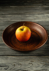 A single bright red apple sits on a brown, patterned wooden plate. The background features a textured wooden table, enhancing the natural look of the setting