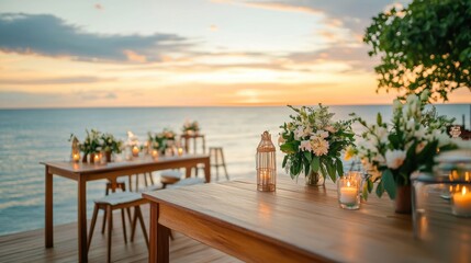A beautifully arranged outdoor dining scene with wooden tables, flowers, candles, and a serene ocean sunset in the background.