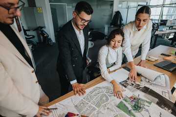 Diverse team gathers around a table to review architectural project plans. The project leader,...
