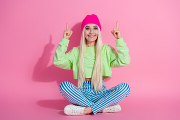 Smiling young woman in colorful casual outfit gesturing hand signs while sitting on the floor against a pink background.