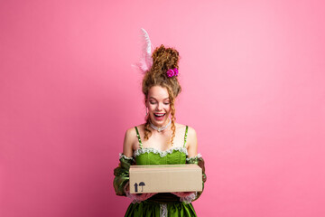 Young woman in a green baroque-inspired dress surprisedly opening a package against a vibrant pink...