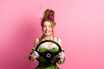 Young woman in retro green gown holding steering wheel with a pink background showcasing an amusing...