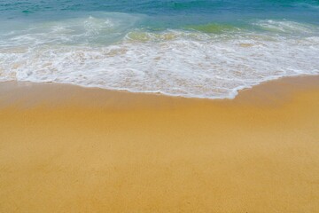 The image shows a sandy beach with gentle waves in the background. The sand is light golden in color and the water has a beautiful blue-green hue. It is a peaceful and relaxing scene.
