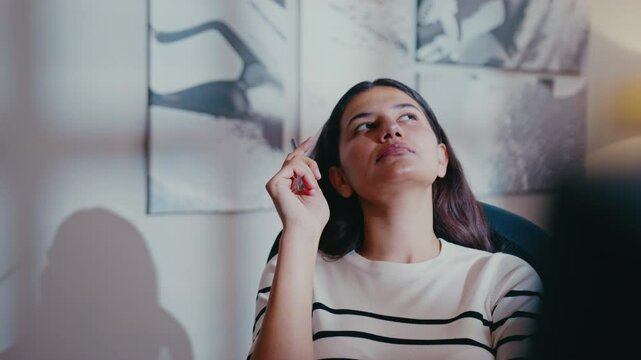 Inspired brunette woman sitting in creative workspace and looking up while holding pen. Thinking deeply and coming up with new concept. Leaning back while brainstorming fresh ideas.